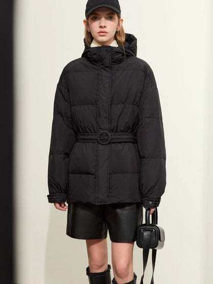 Model stands facing forward wearing a black puffer jacket with a high neck and hood. The jacket features a belt at the waist for a cinched silhouette. She is styled with black leather shorts, black knee-high boots, a black baseball cap, and a black mini handbag