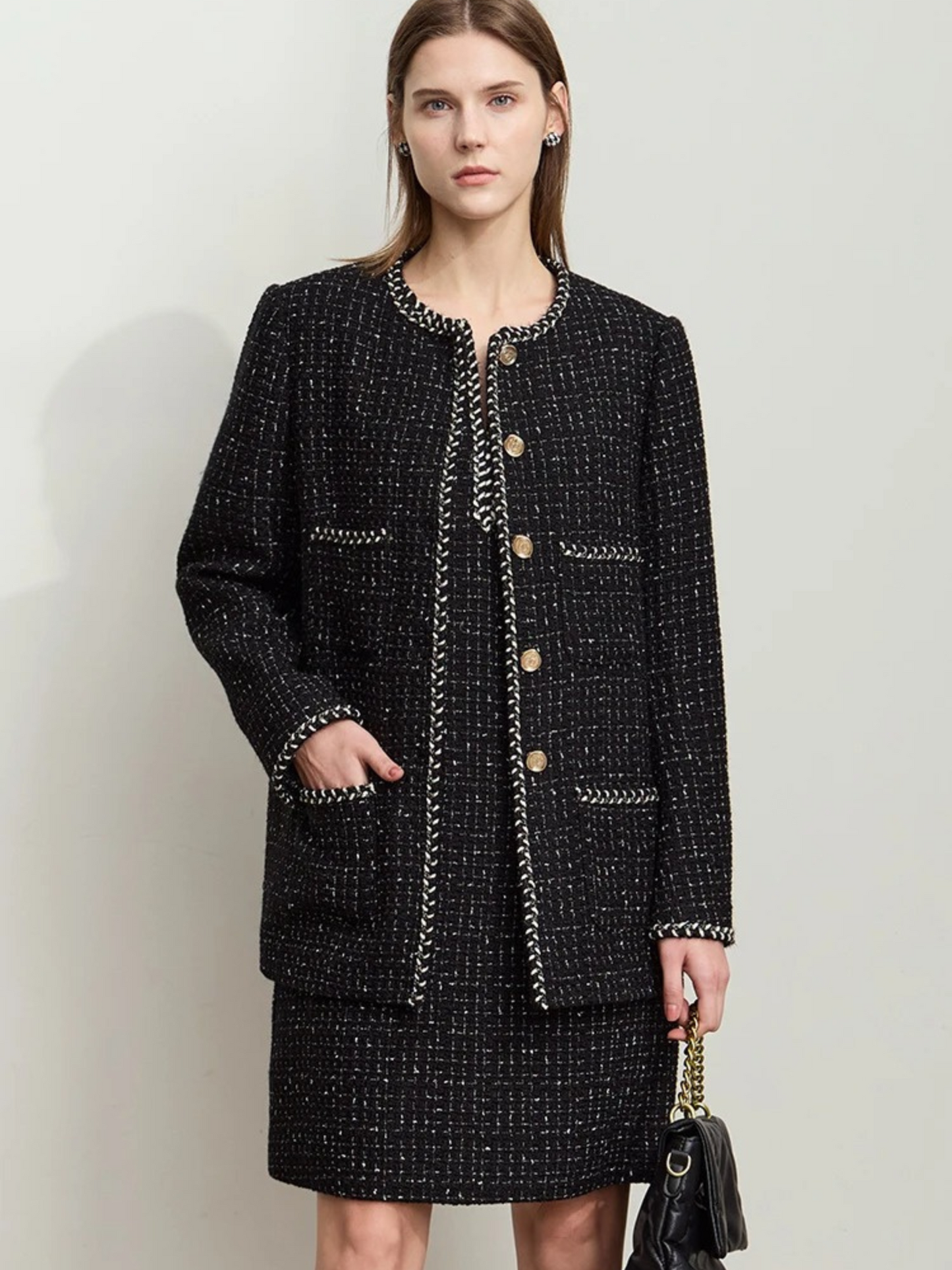 A female model stands facing forward, wearing a textured black tweed jacket with gold buttons and a matching black skirt. She holds a black handbag in her left hand. The jacket has a round neckline, decorative pockets, and white trim along the edges