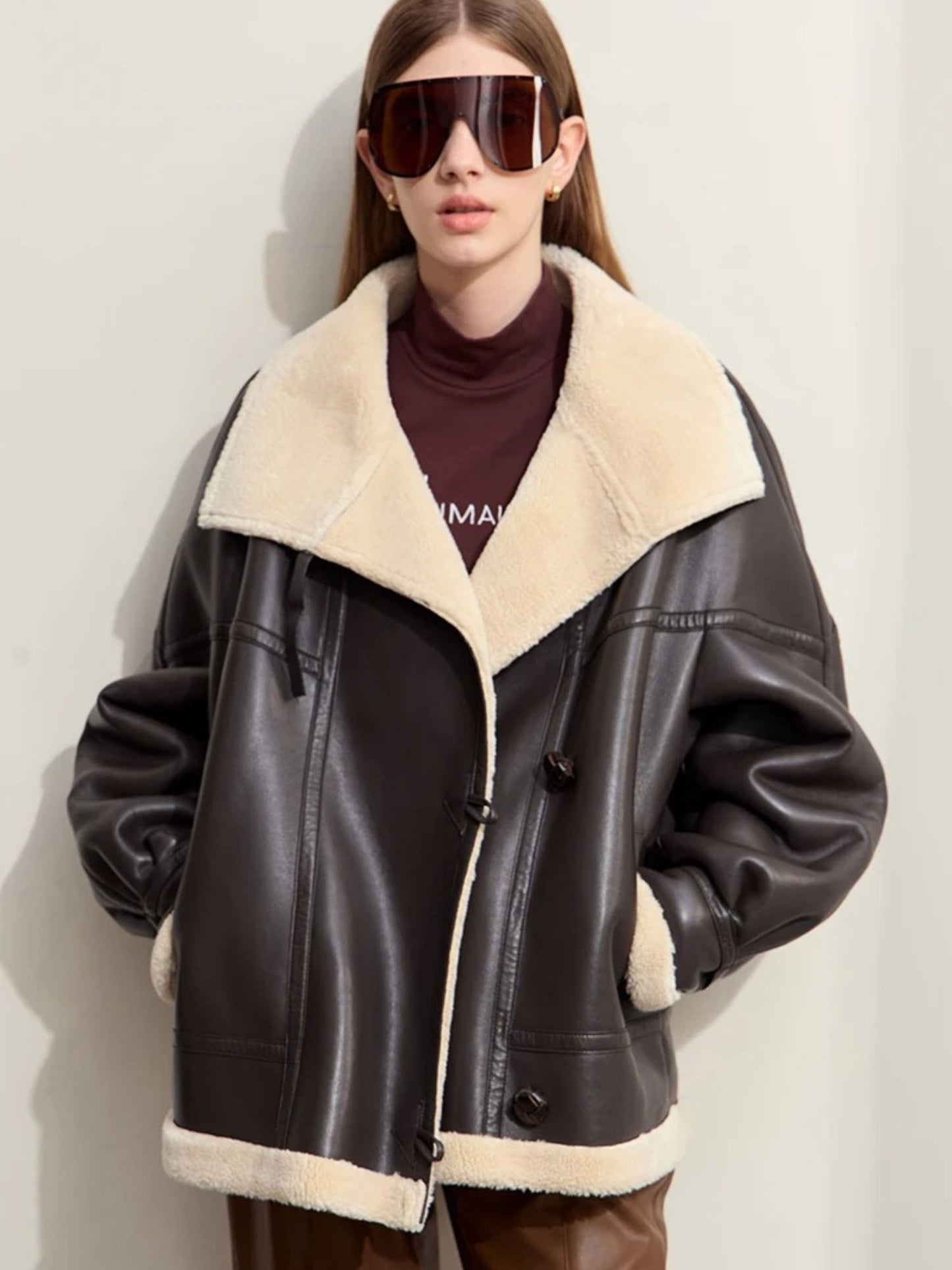 A female model stands facing forward, wearing a black faux leather oversized aviator jacket with a cream shearling lining and collar. The jacket features silver zipper details and large lapels. She wears brown pants, a burgundy turtleneck, and oversized sunglasses