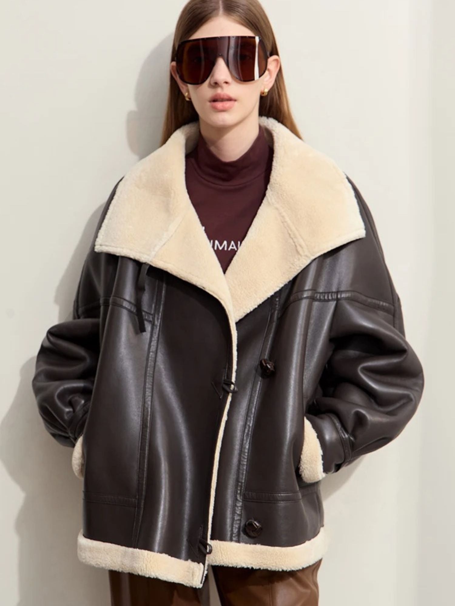 A female model stands facing forward, wearing a black faux leather oversized aviator jacket with a cream shearling lining and collar. The jacket features silver zipper details and large lapels. She wears brown pants, a burgundy turtleneck, and oversized sunglasses