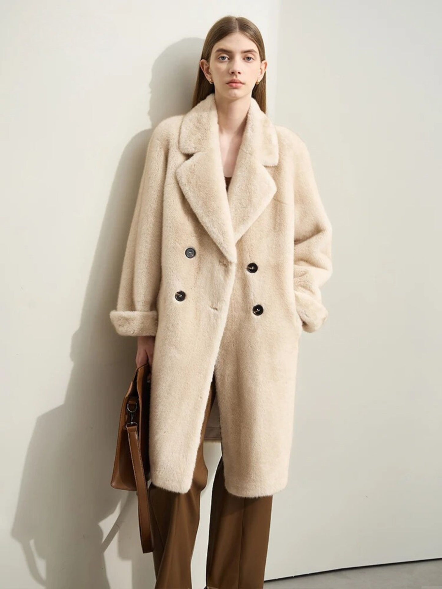 A female model stands front-facing, leaning against a light wall, wearing a cream-colored wool coat with an oversized collar and double-breasted button closure. The coat falls to the knees and has a slightly textured surface. She pairs it with brown wide-leg trousers and holds a brown handbag