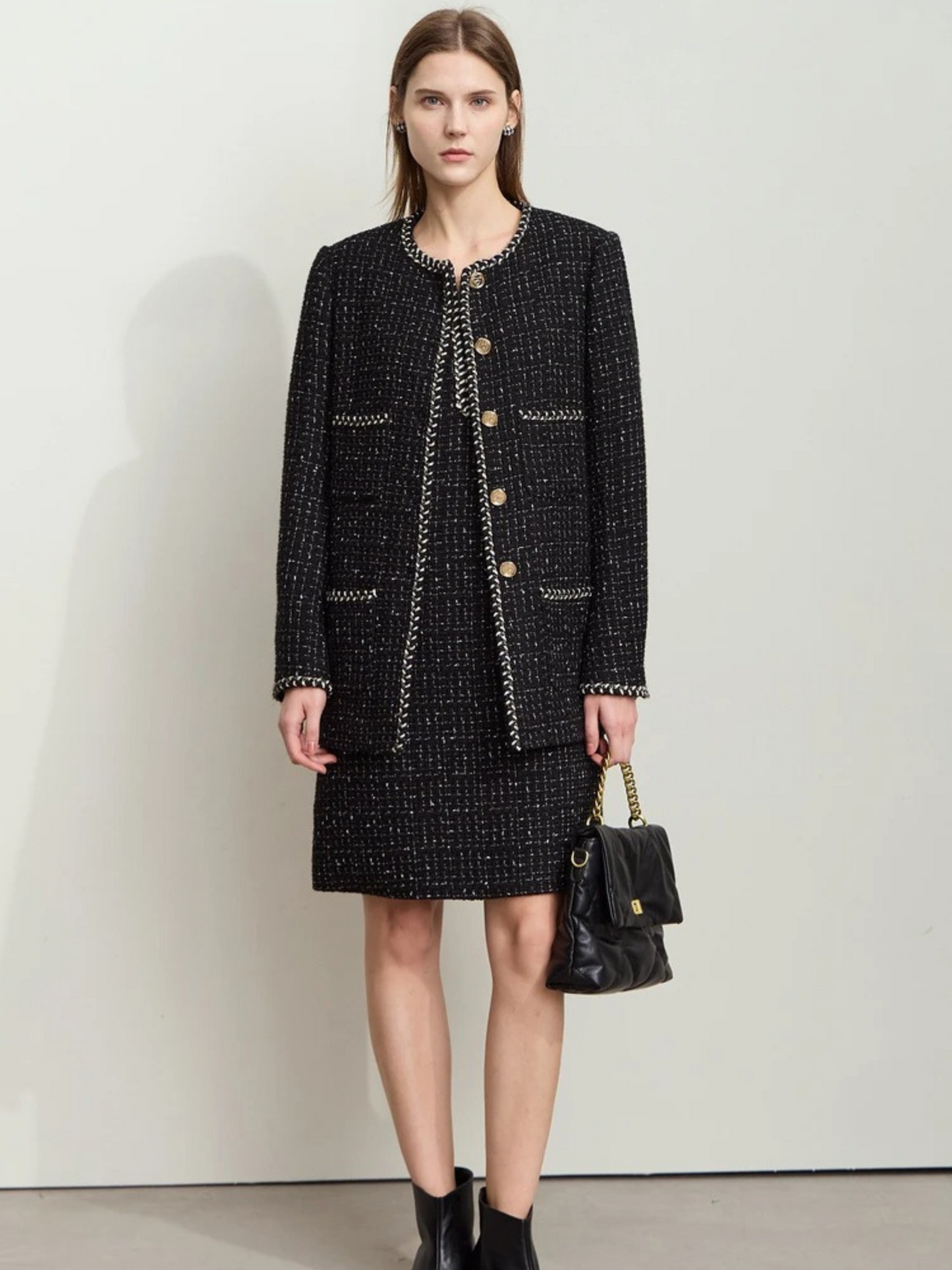 Full-body shot of the same model wearing the black tweed outfit, standing against a plain white wall. She holds the handbag with both hands in front