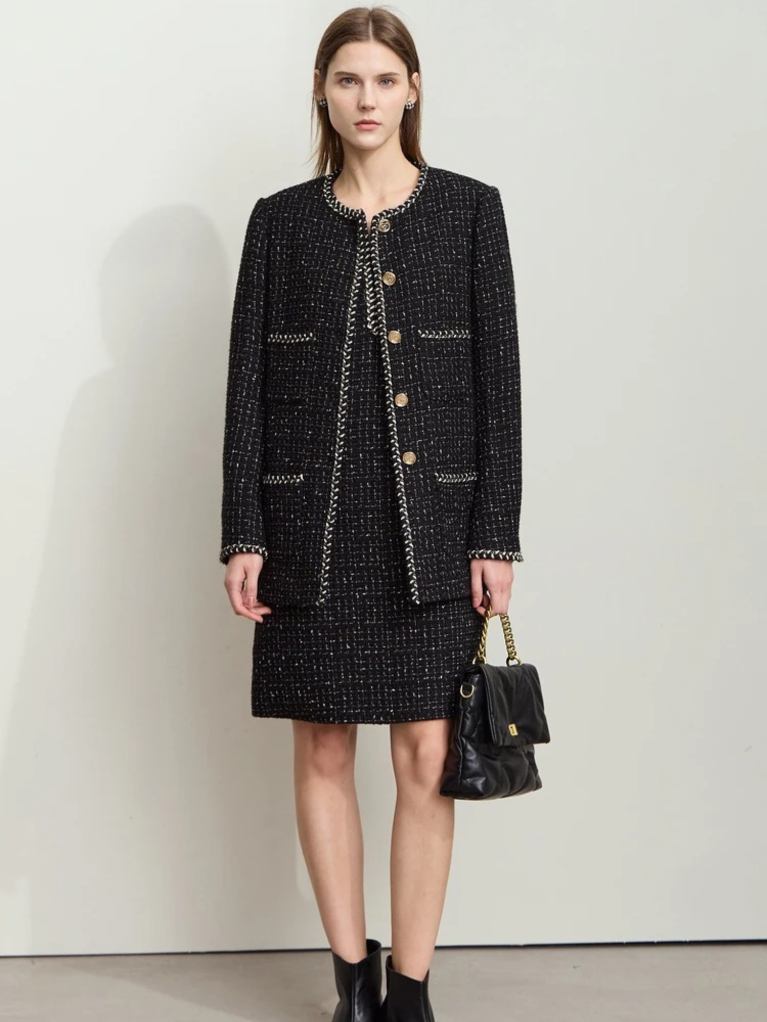 Full-body shot of the same model wearing the black tweed outfit, standing against a plain white wall. She holds the handbag with both hands in front