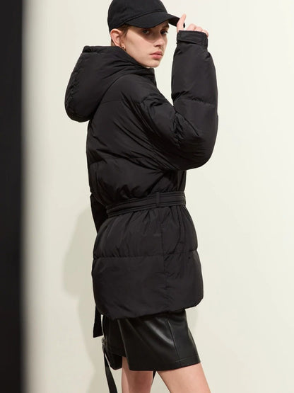 Side profile of the model in the black puffer jacket. She adjusts her black cap while holding the same mini handbag. The jacket appears slightly puffed and insulated