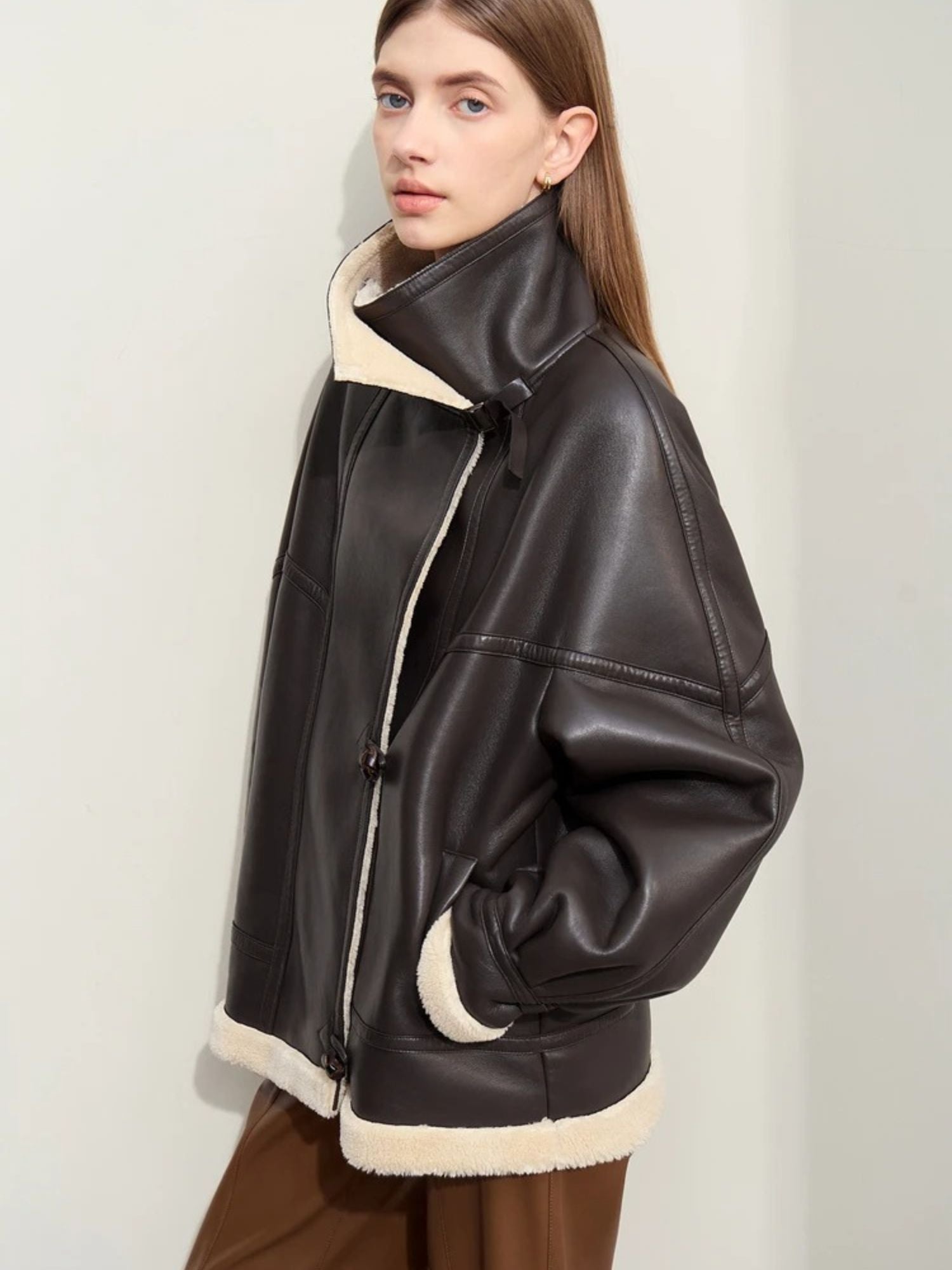 Side profile view of the model showing the oversized silhouette of the jacket. She holds her hands casually in the jacket’s side pockets, and the shearling lining is visible at the collar and hem