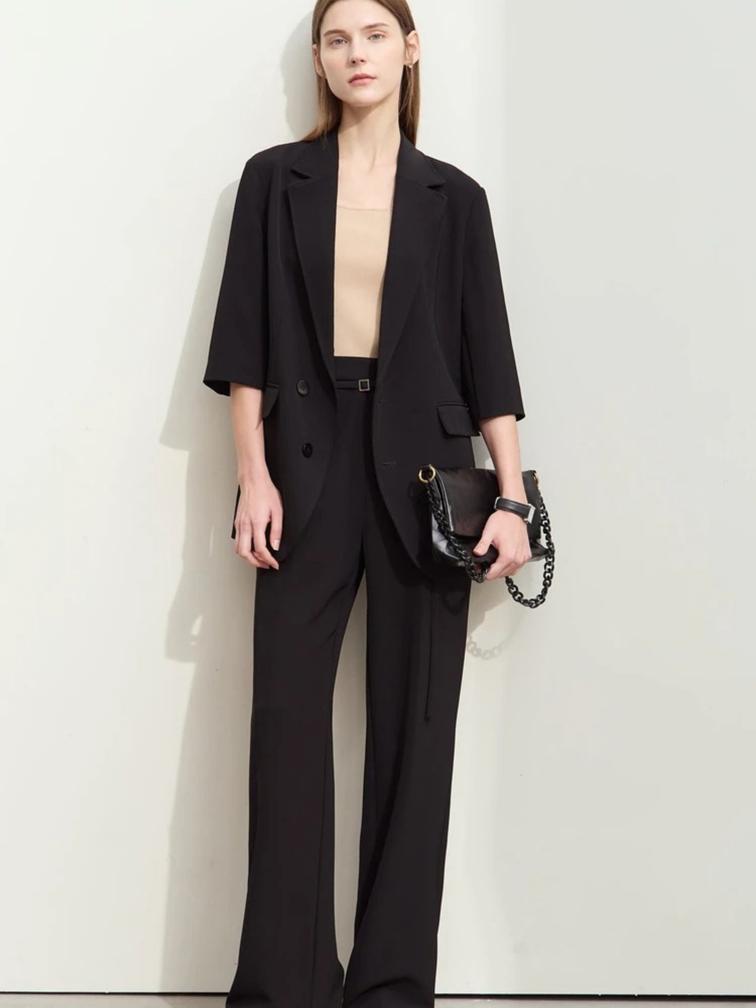 Front-facing woman in black wide-leg pants and elbow-sleeve blazer over a beige top, holding a black handbag