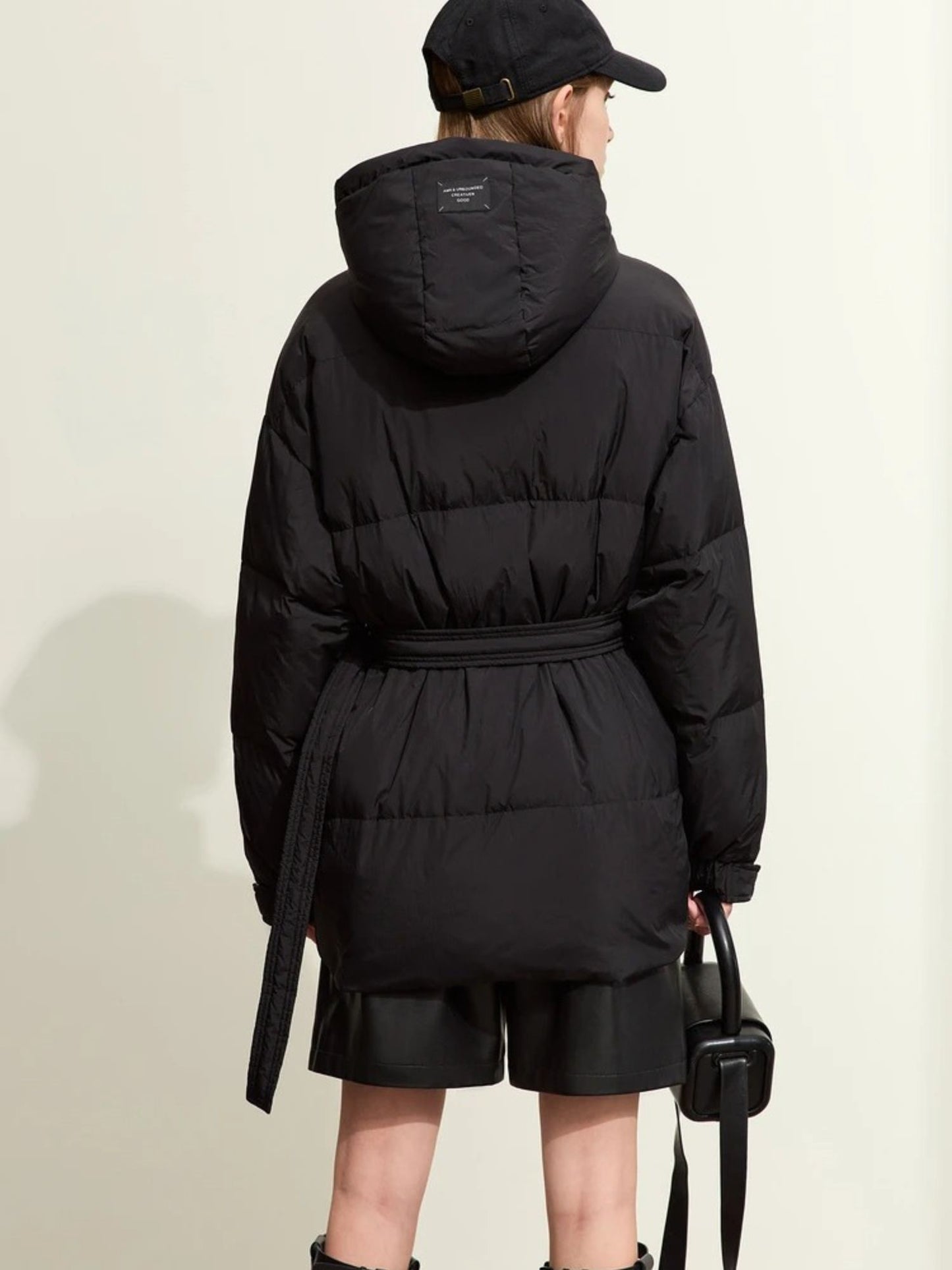 Back view of the black puffer jacket, showing the hood, horizontal quilted sections, and belt detail at the back. The model’s arms hang naturally