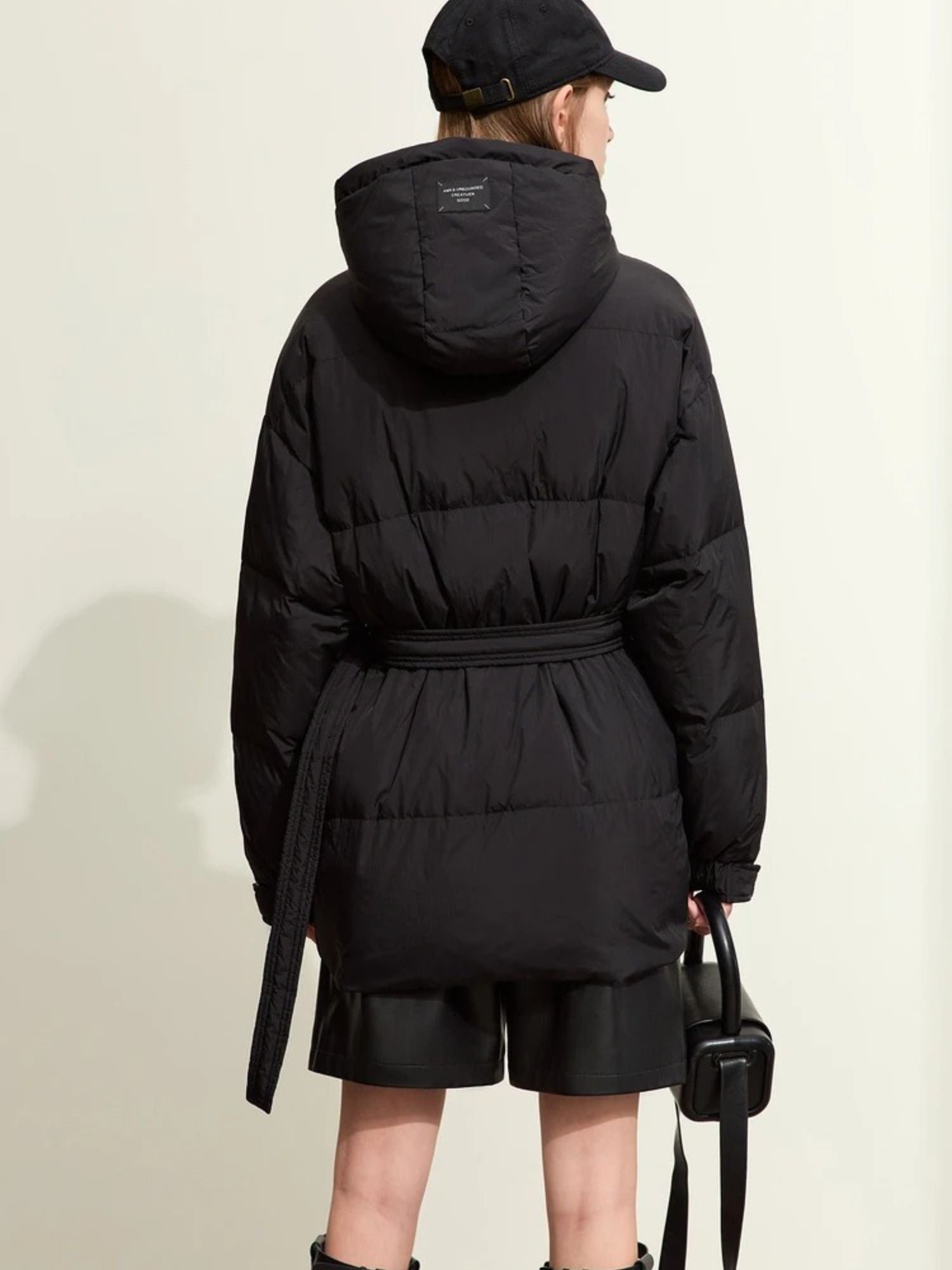 Back view of the black puffer jacket, showing the hood, horizontal quilted sections, and belt detail at the back. The model’s arms hang naturally