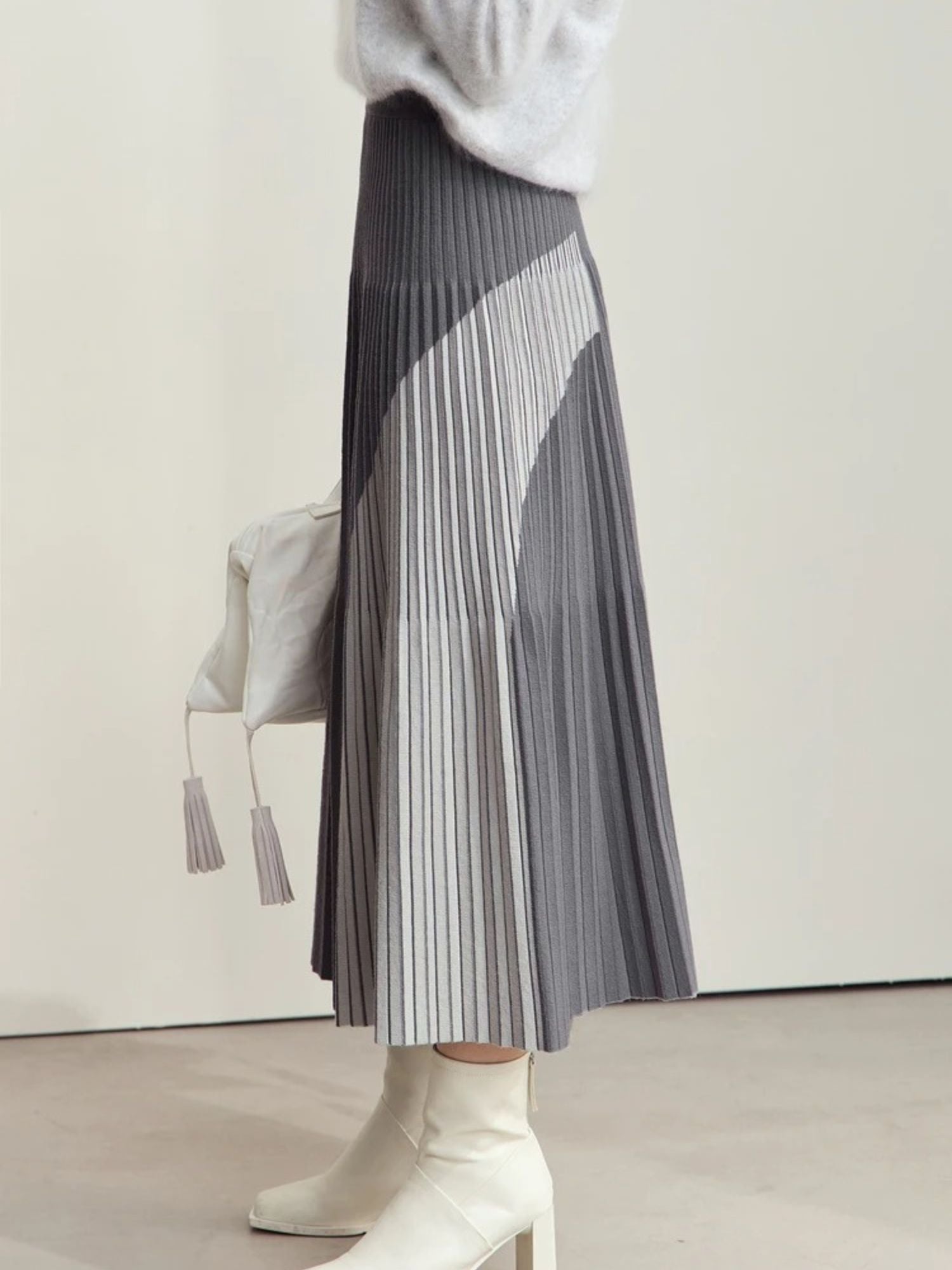Model walking in gray pleated midi skirt with color-block panels, paired with gray sweater and white boots