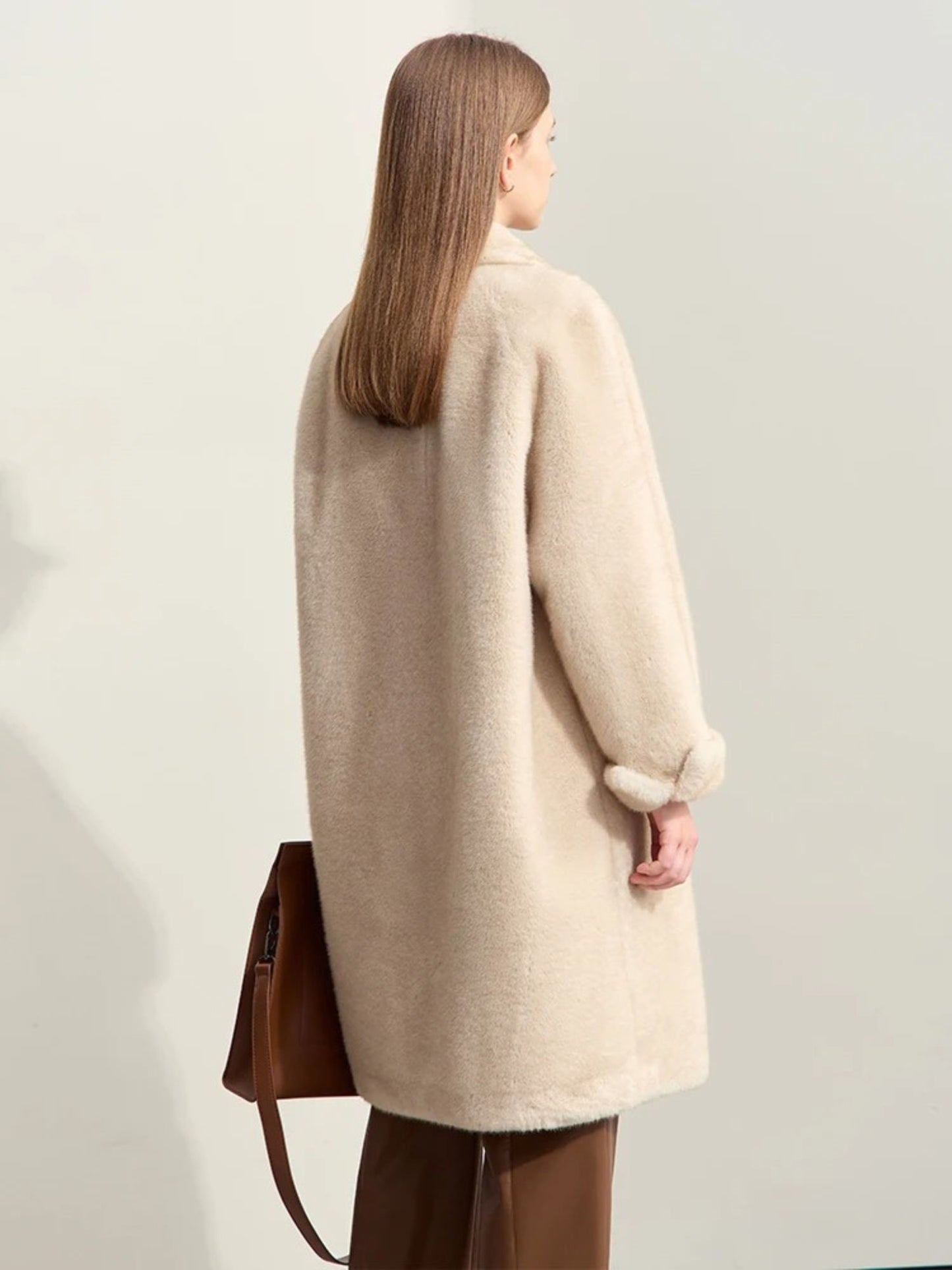 Rear view of the coat, showing its relaxed fit and straight back seam. The coat drapes loosely, and the model’s brown handbag is visible hanging from her shoulder