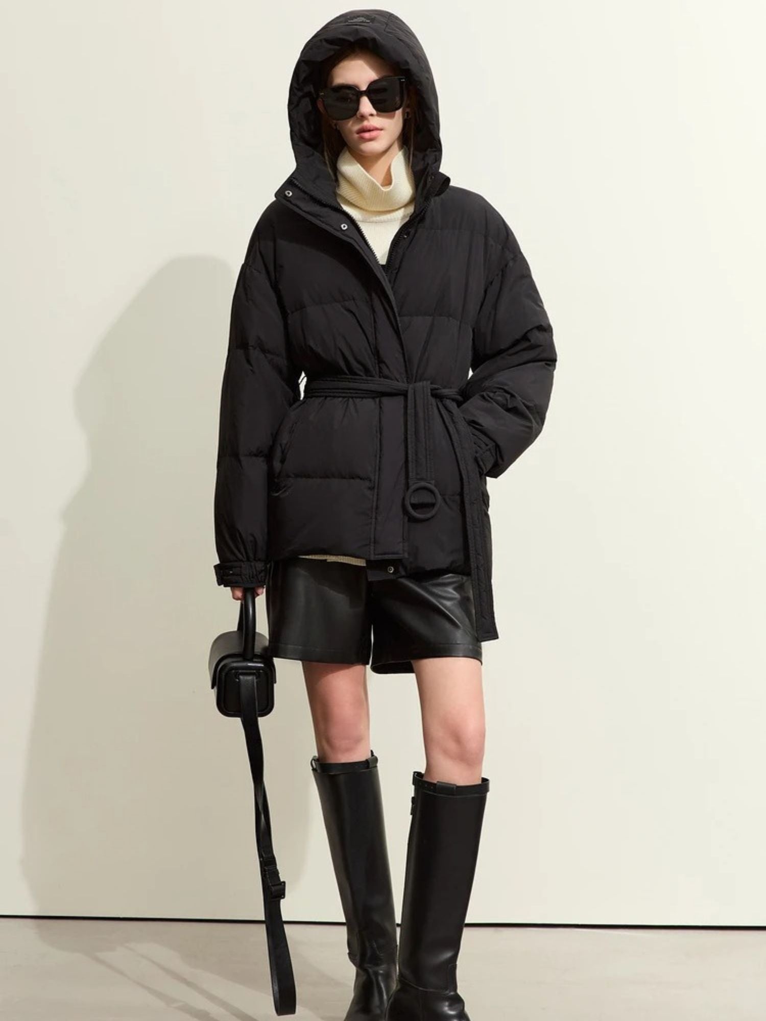 Front view of the model wearing the black puffer with a black cap and knee-high boots, standing in a neutral pose. Her hands are by her sides, showing the structure of the jacket clearly