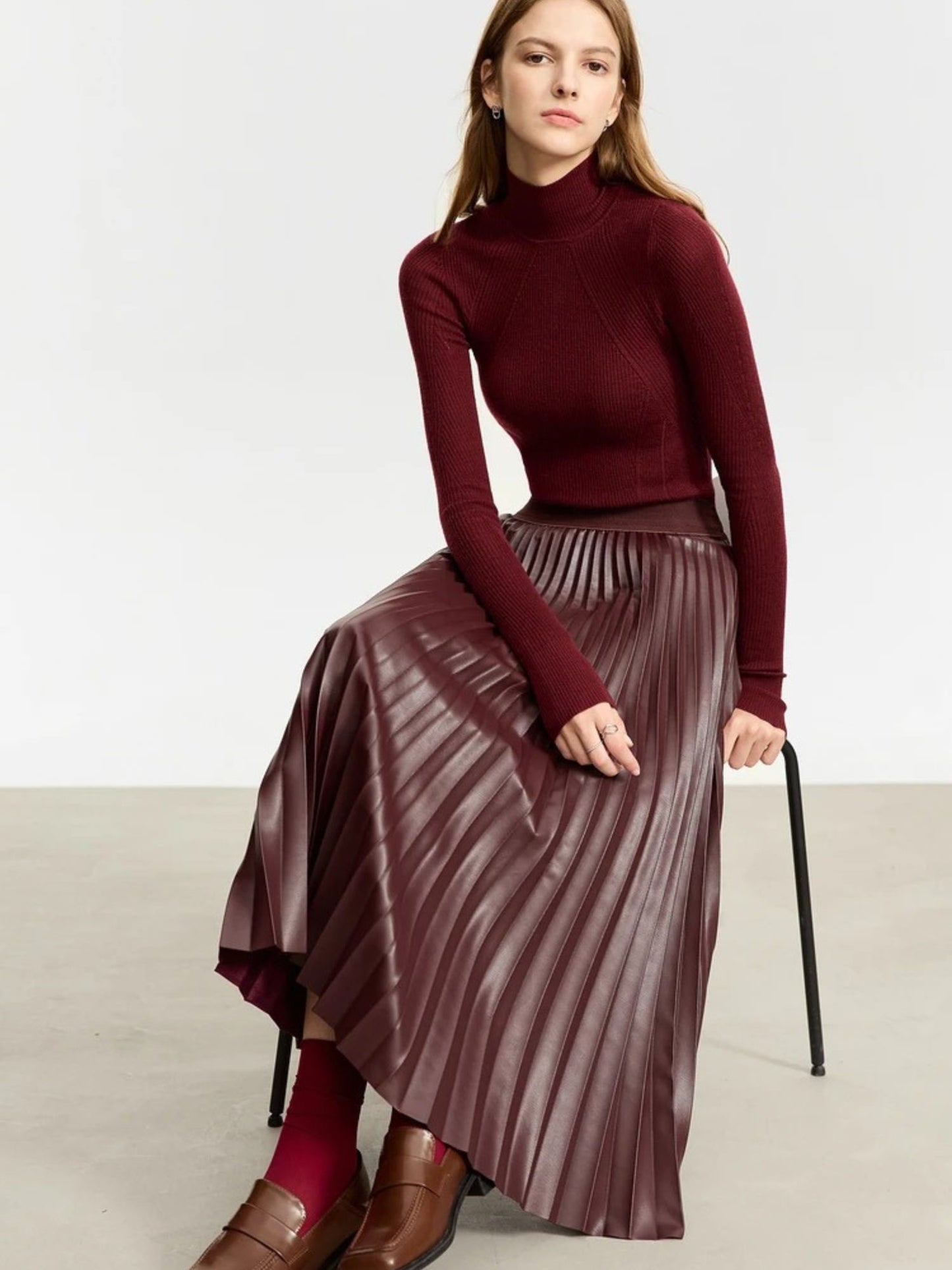 Seated front view of burgundy faux leather pleated midi skirt paired with matching burgundy top