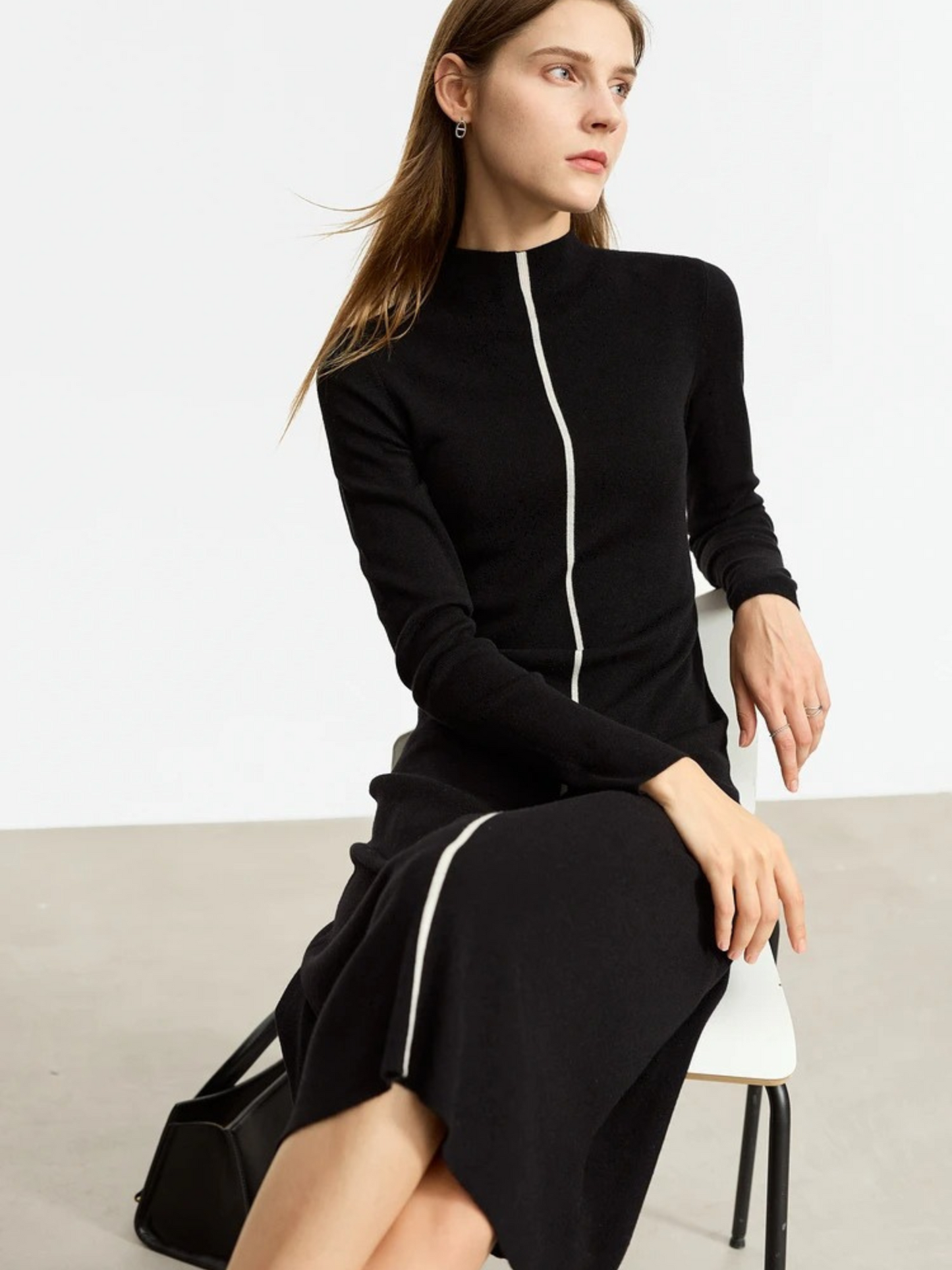 Seated model wearing black long-sleeve midi dress with front zipper, fitted silhouette, styled with black boots