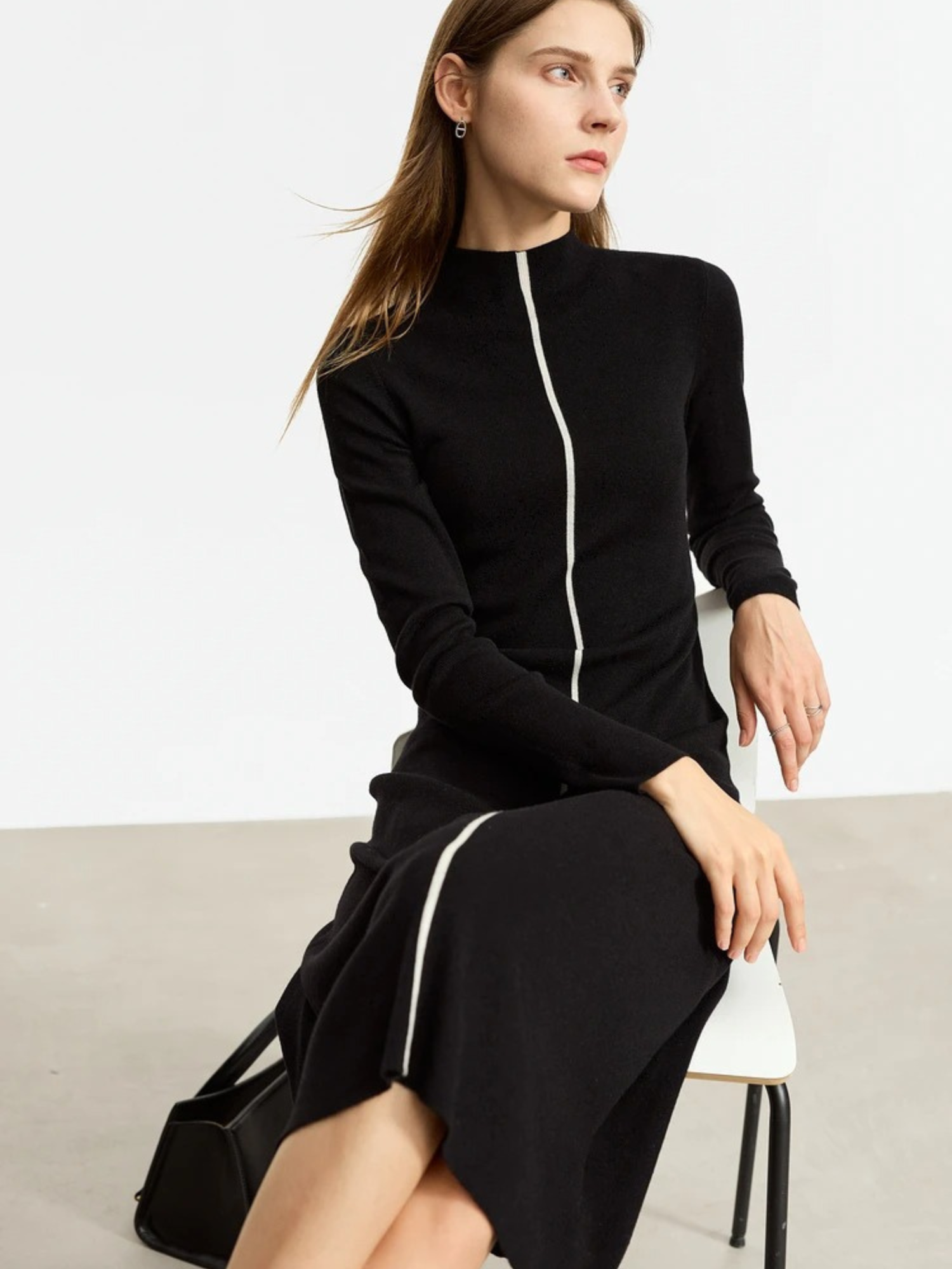 Seated model wearing black long-sleeve midi dress with front zipper, fitted silhouette, styled with black boots