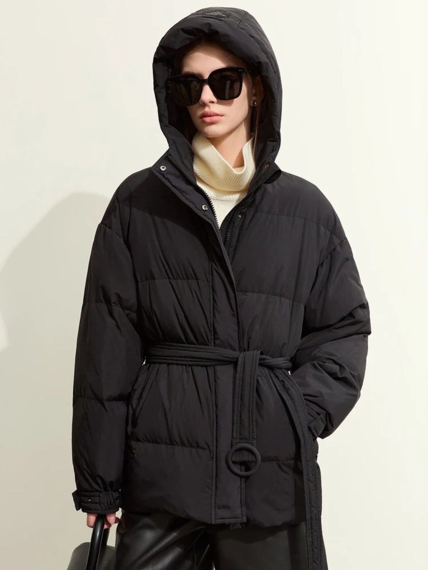 The same black puffer jacket, but styled with black sunglasses and without a cap. The model’s hands are in her pockets. The sunlight casts a warm glow, enhancing the texture of the jacket